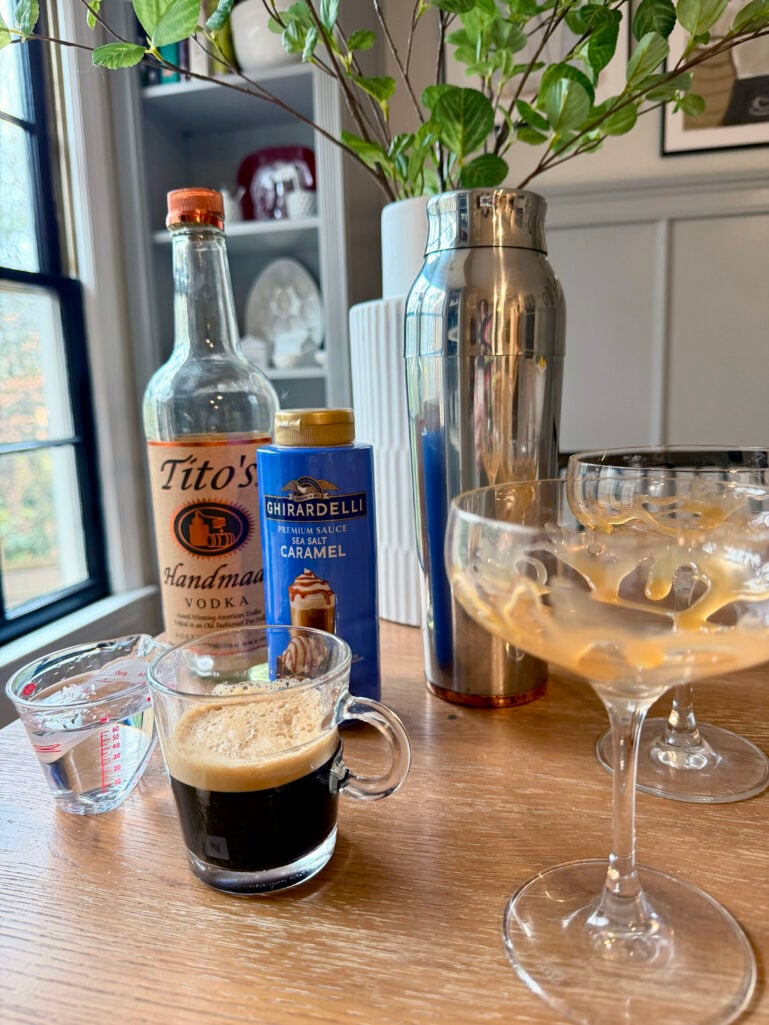 Salted Caramel Espresso Martini ingredients arranged on a wooden table: Tito's vodka, Ghirardelli sea salt caramel sauce, fresh espresso, a cocktail shaker, and coupe glasses drizzled with caramel