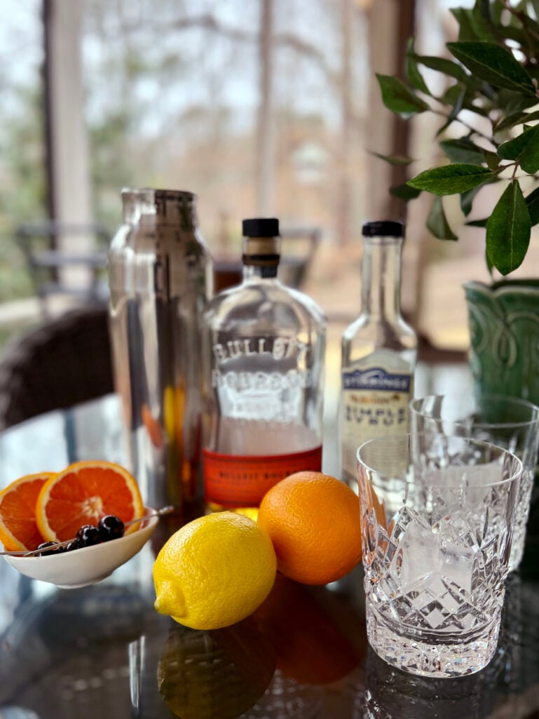 Blood orange whiskey sour ingredients including Bulleit Bourbon, blood oranges, lemon, simple syrup, brandied cherries, cocktail shaker, and crystal rocks glasses with ice