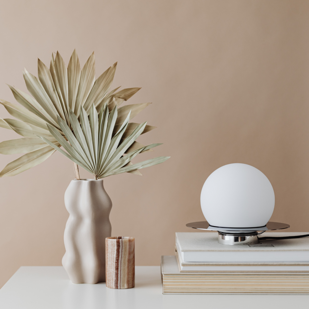 Minimalist tabletop styling featuring a sculptural cream ceramic vase with dried palm leaves, a small stone. candle holder, stacked neutral books, and a modern globe table lamp against a warm beige backdrop