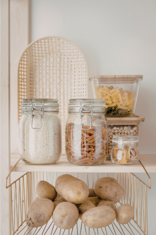 Open pantry shelving with clear glass storage jars filled with rice, pasta, and dried goods, stacked glass containers with pasta, and a wire basket holding potatoes below for organized kitchen storage.