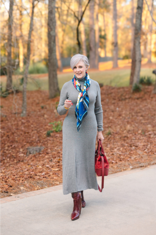 Woman wearing a gray ribbed sweater dress styled with a colorful silk scarf, burgundy leather boots, and a matching red handbag, walking along a paved path with autumn leaves and trees in the background.