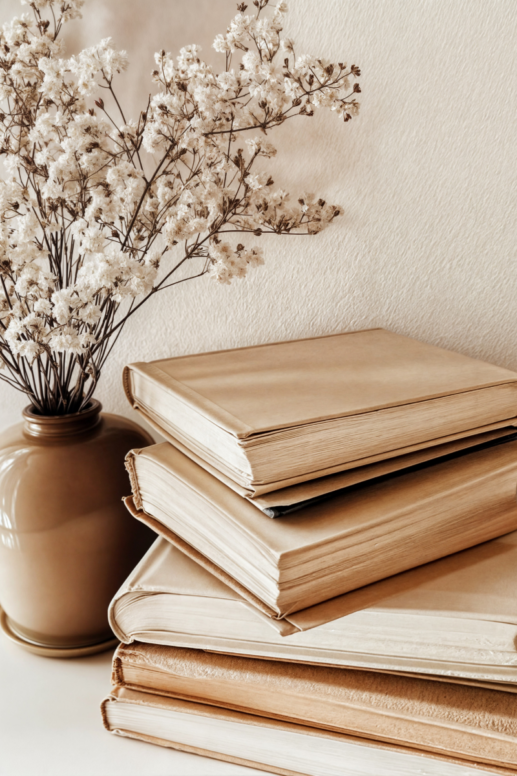 Neutral-toned still life featuring a stack of vintage-style books with soft beige covers beside a ceramic vase filled with dried white flowers, creating a calm, minimalist home decor vignette.