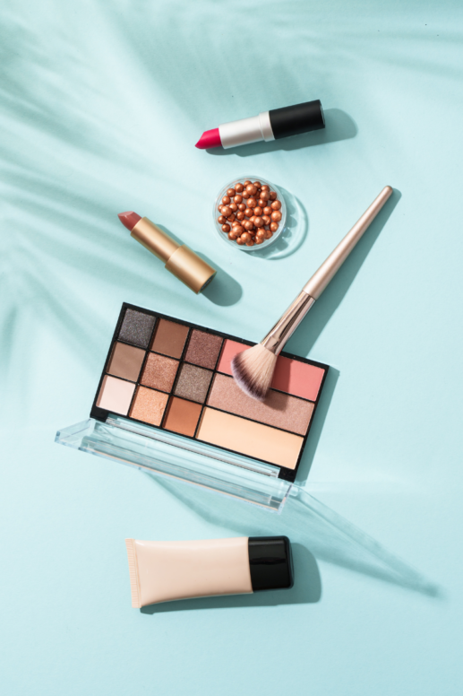Flat lay of makeup products on a soft blue background, including an eyeshadow palette with neutral and rosy shades, a blush brush, two lipsticks in pink and nude tones, bronzing pearls in a clear jar, and a tube of foundation, styled in natural light with soft shadows.