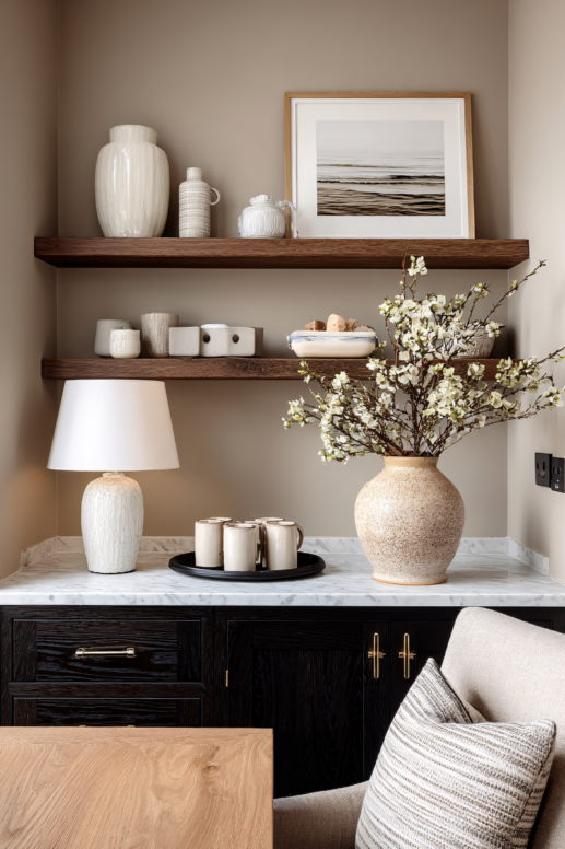 Styled home interior featuring dark wood cabinetry topped with a marble surface, layered wooden shelves with neutral ceramics and framed art, a textured table lamp, and a large stone vase filled with white flowering branches for a warm, modern aesthetic.