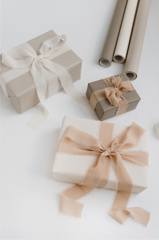 Neutral-toned gift boxes wrapped in soft beige and ivory paper with frayed silk ribbons, styled alongside rolled wrapping paper for an elegant, minimalist gift presentation.