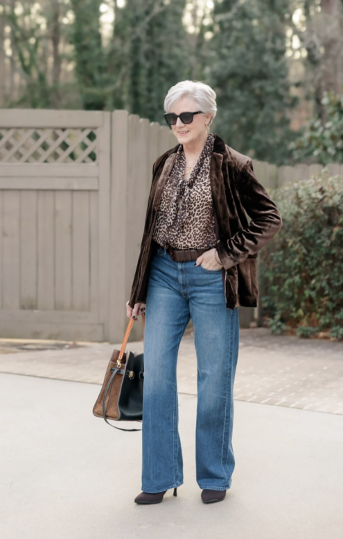 Woman wearing a brown velvet blazer, leopard-print blouse, and wide-leg blue jeans, styled with heeled boots and a structured handbag, walking outdoors along a paved path.