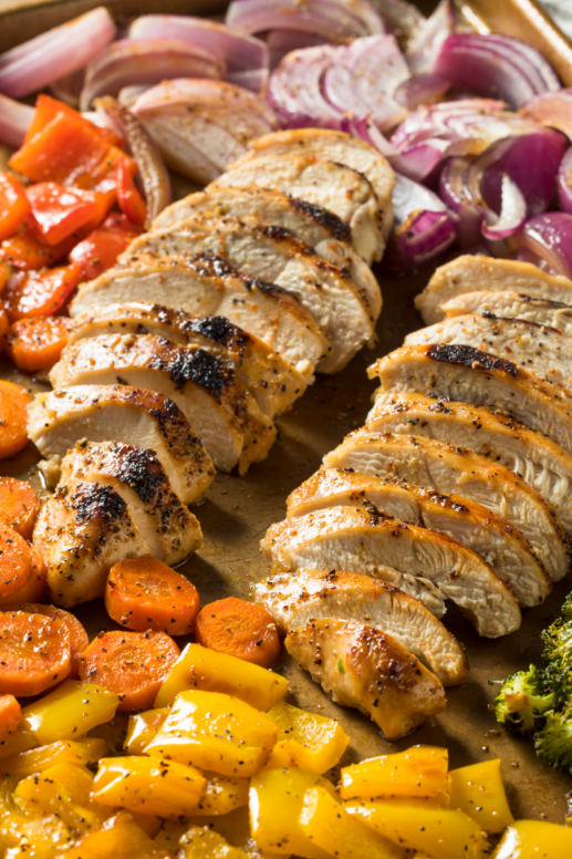 Sheet-pan dinner with sliced roasted chicken breast surrounded by colorful vegetables, including carrots, bell peppers, red onions, and broccoli, seasoned and roasted until golden.