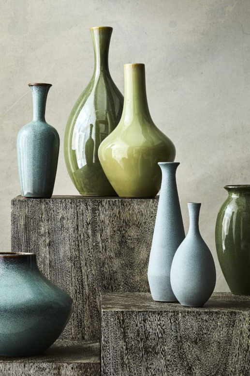Collection of glazed ceramic vases in soft blue and green tones, arranged on rustic wooden pedestals to create a layered, minimalist home decor display.