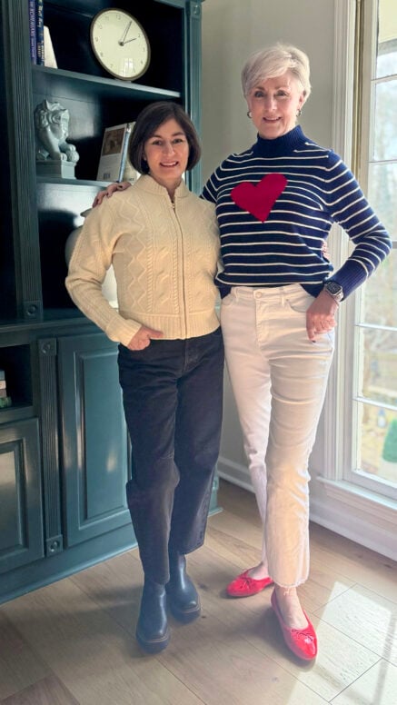 Beth and Kelly standing together in front of teal built-in bookshelves, Beth in a navy striped sweater with a red heart and Kelly in a cream cable knit bomber jacket