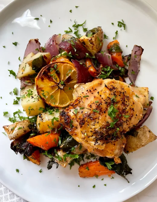 Plated lemon pepper chicken thigh with crispy golden skin on a bed of roasted potatoes, carrots, red onion, and crispy spinach, topped with a caramelized lemon wheel and fresh parsley on a white plate