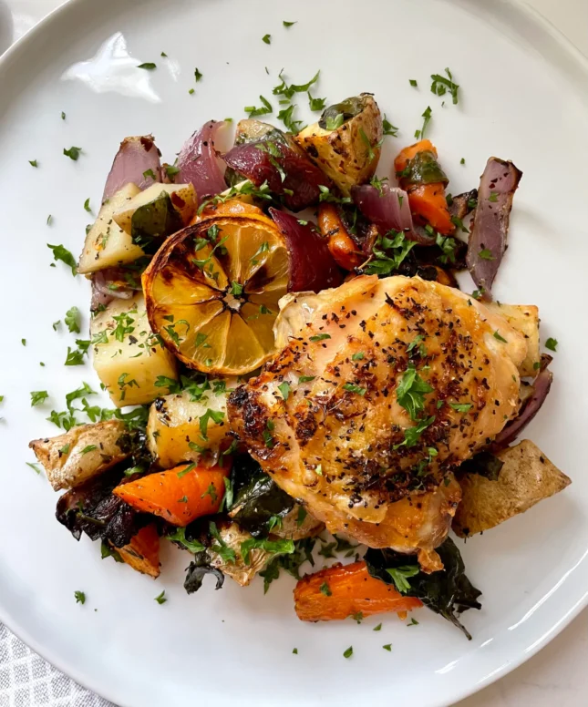 Plated lemon pepper chicken thigh with crispy golden skin on a bed of roasted potatoes, carrots, red onion, and crispy spinach, topped with a caramelized lemon wheel and fresh parsley on a white plate