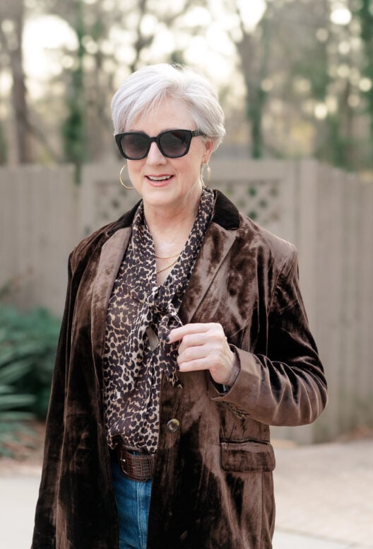Beth wearing chocolate brown velvet blazer over leopard print tie-neck blouse with gold hoop earrings, layered gold necklace, and black sunglasses