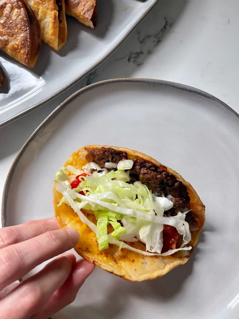 Hand holding an open crispy taco filled with beef, lettuce, tomato, cheese, and sour cream