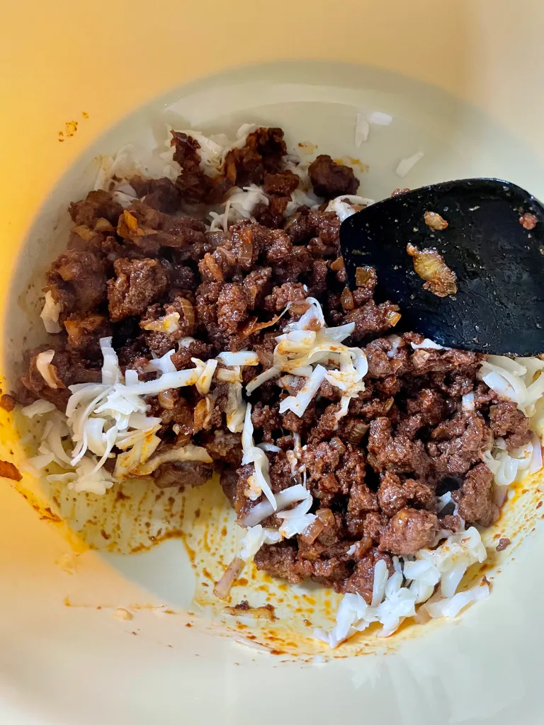 Cooked taco meat being mixed with shredded cheddar cheese in a bowl