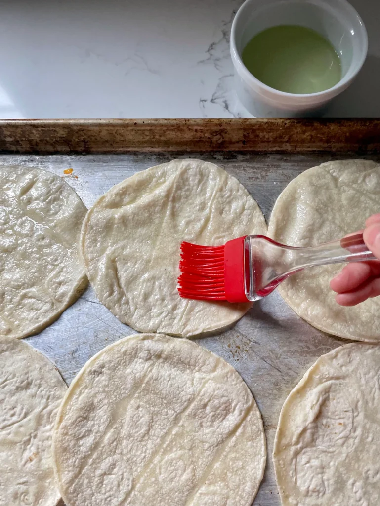 Brushing corn tortillas with oil on a sheet pan using a red silicone brush