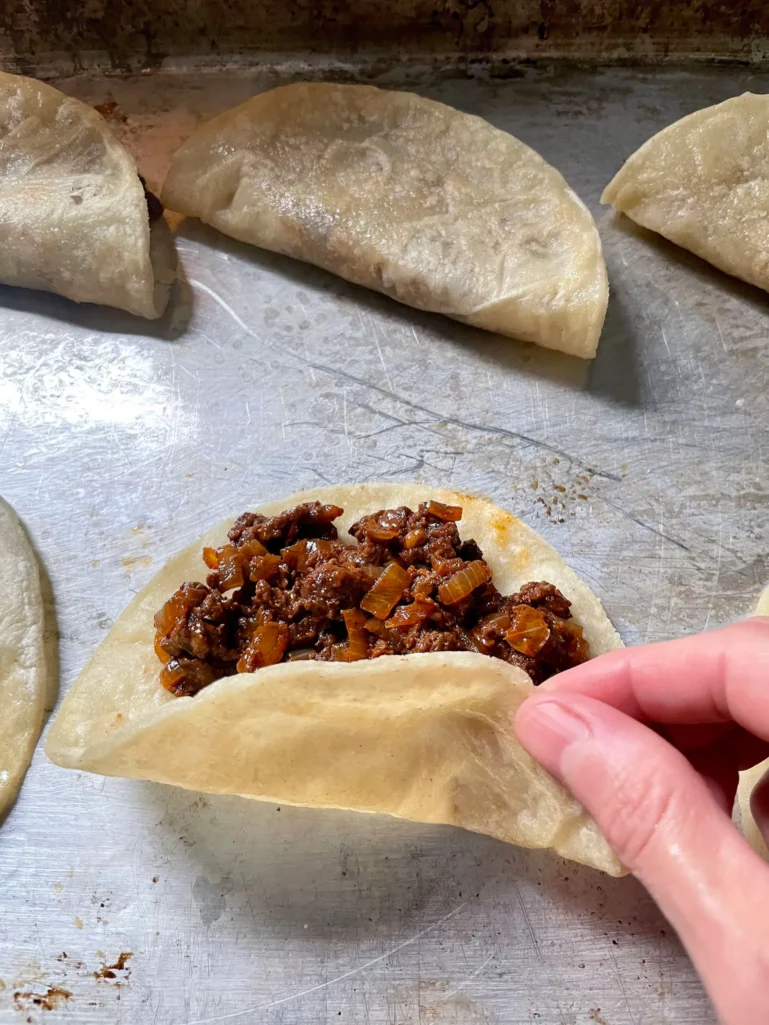 Filling a warmed corn tortilla with seasoned beef on a sheet pan with folded tacos in background
