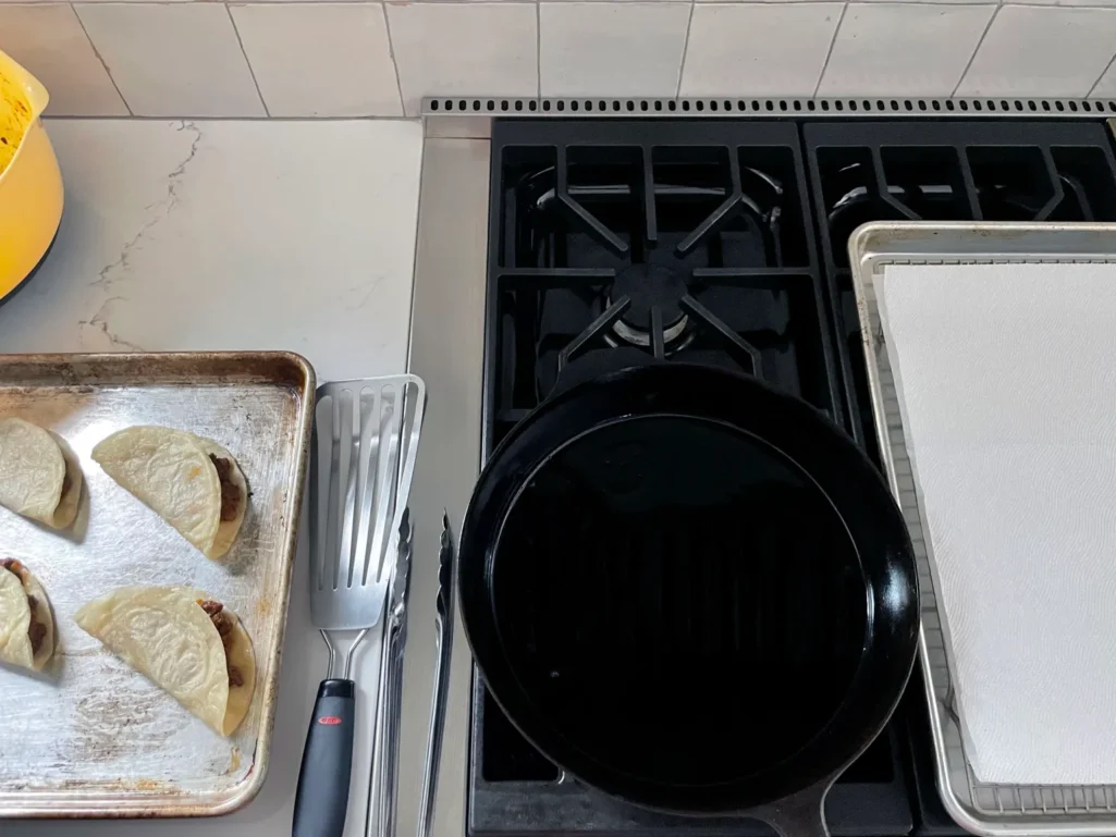 Overhead view of taco frying station setup with filled tacos on sheet pan, cast iron skillet, spatula, tongs, and paper towel-lined sheet pan