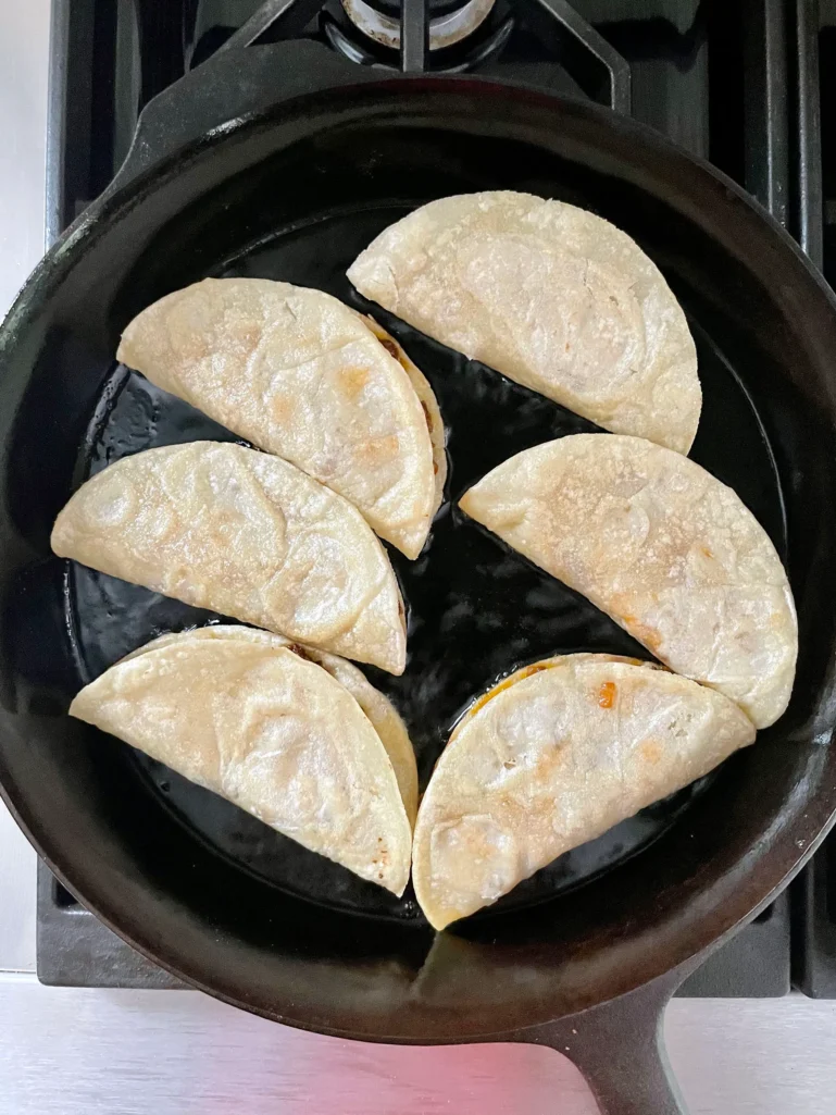 Six filled tacos starting to fry in oil in a cast iron skillet