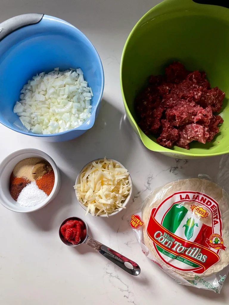 Overhead view of tacos dorados ingredients: chopped onion, ground beef, spices, shredded cheese, tomato paste, and corn tortillas