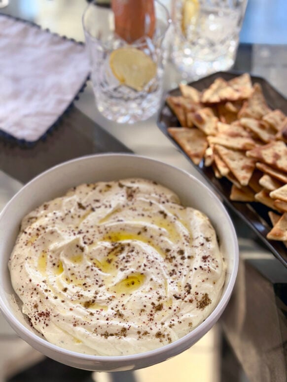 Creamy whipped feta swirled in a white bowl, drizzled with olive oil and sprinkled with za'atar, served with pita chips and mocktails in the background
