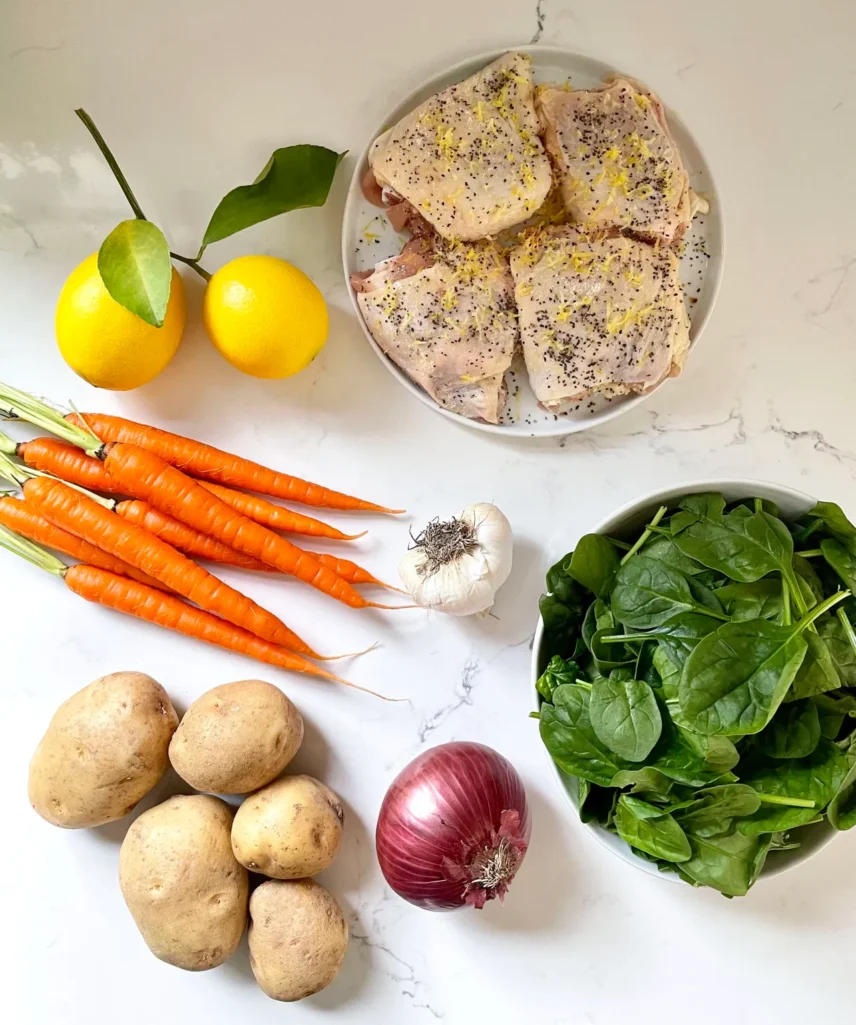 Overhead view of sheet pan chicken ingredients: seasoned chicken thighs with lemon zest and pepper, fresh lemons, whole carrots, garlic, potatoes, red onion, and fresh spinach in a bowl on a marble countertop