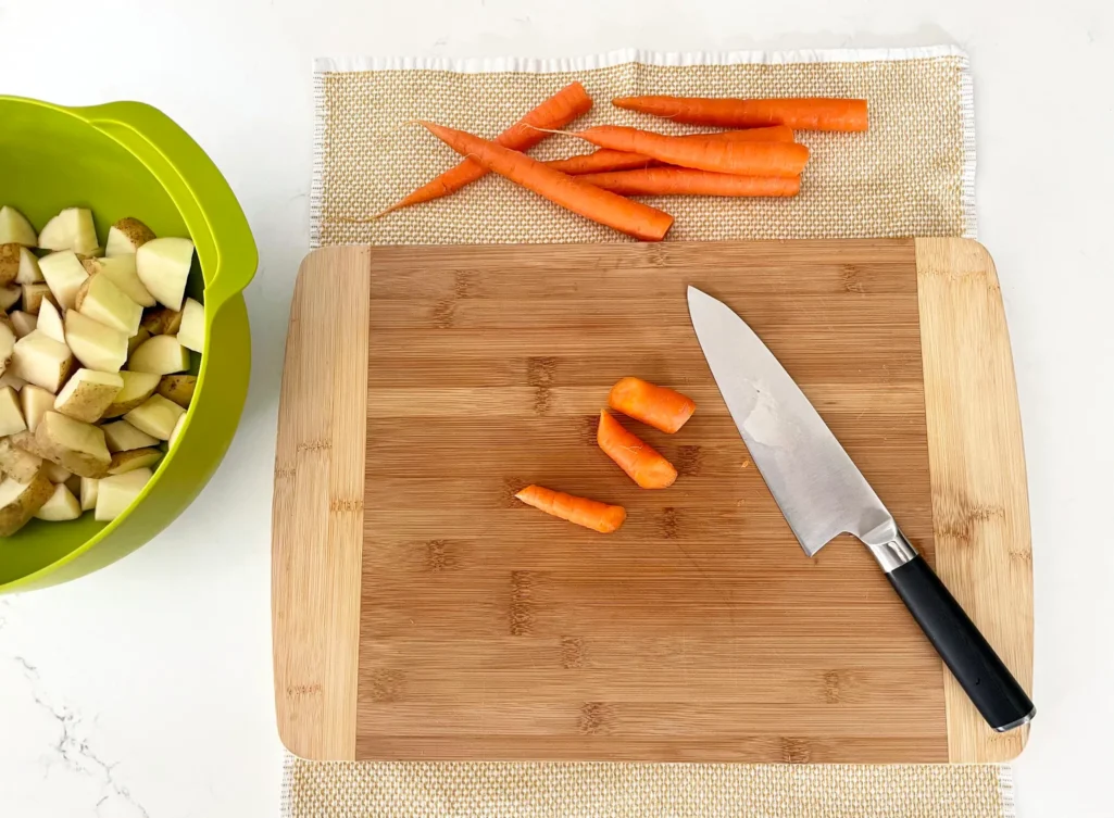 Veggie prep station with bamboo cutting board on a dish towel, carrots being chopped, and a green bowl of cubed potatoes