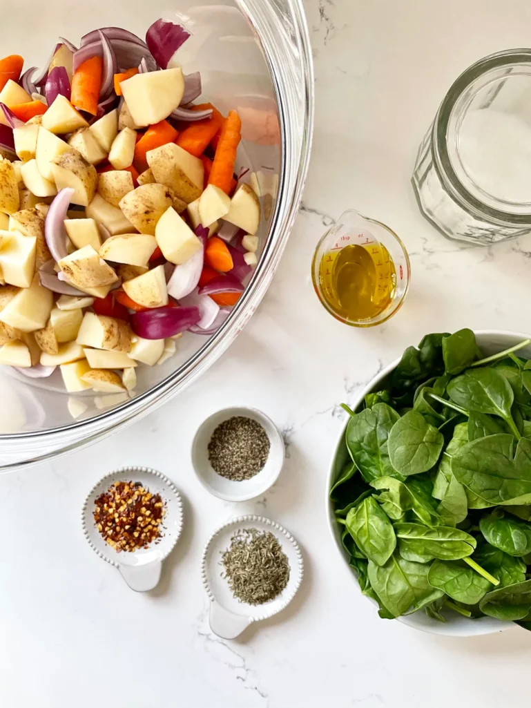 Chopped potatoes, carrots, and red onion in a large glass bowl with small dishes of red pepper flakes, dried thyme, and herbs, olive oil in a measuring cup, and fresh spinach ready to mix