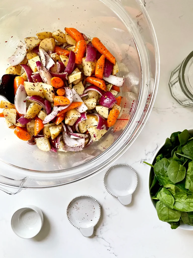 Vegetables tossed with olive oil and seasonings in a large glass Cambro container with fresh spinach in a bowl nearby