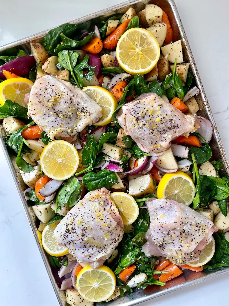 Unbaked sheet pan with raw seasoned chicken thighs arranged over vegetables and spinach, topped with fresh lemon slices before roasting