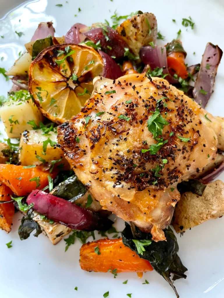 Close-up of crispy-skinned lemon pepper chicken thigh garnished with fresh parsley alongside roasted vegetables and a caramelized lemon wheel