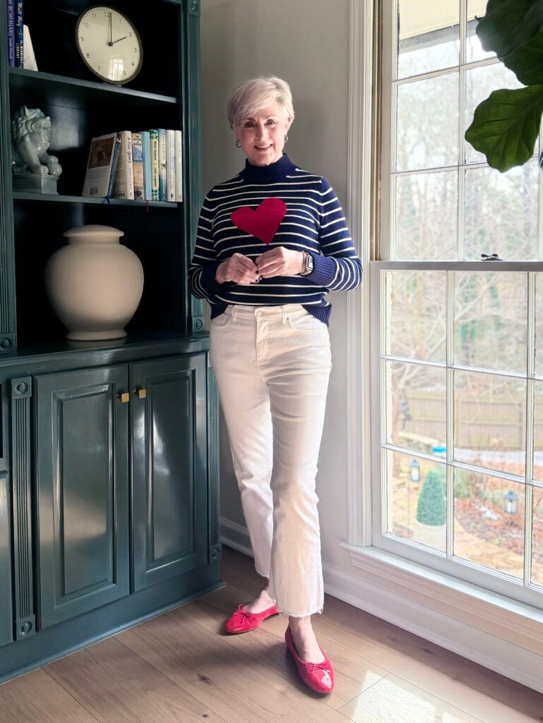 Beth wearing a navy and white striped mockneck sweater with a red heart detail, cream denim, and red ballet flats, standing by the window