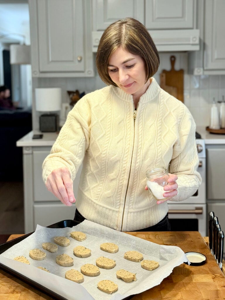 Kelly in her cream cable knit sweater sprinkling flaky sea salt over brown butter shortbread rounds on a parchment-lined baking sheet