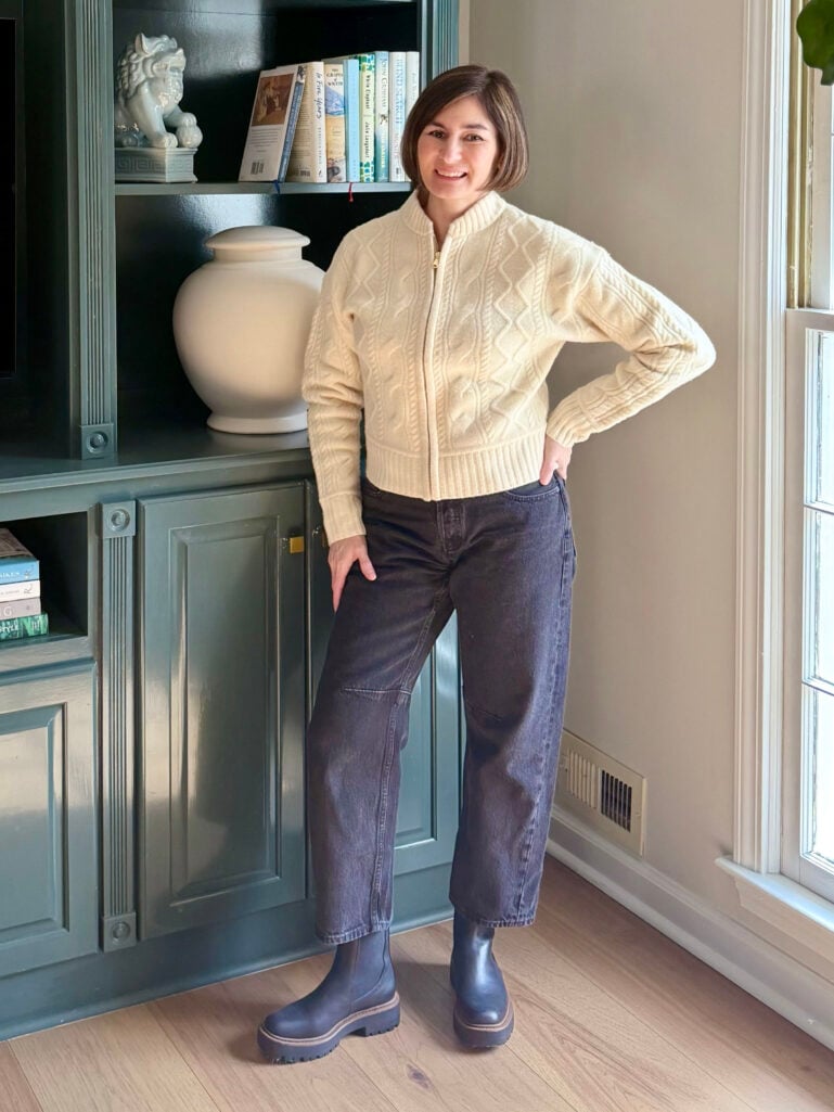 Full-length view of Kelly in her cream bomber and dark jeans, standing in front of teal built-ins with books and decor