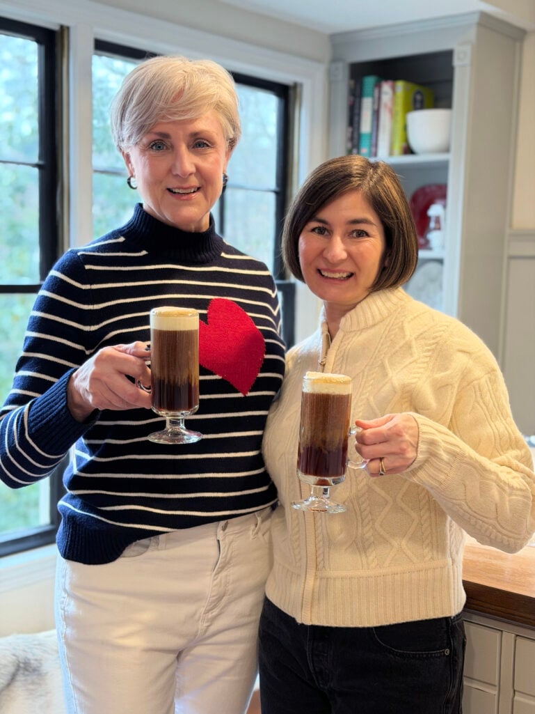 Beth and Kelly holding Irish Coffees topped with cream, smiling at the camera in their cozy sweaters