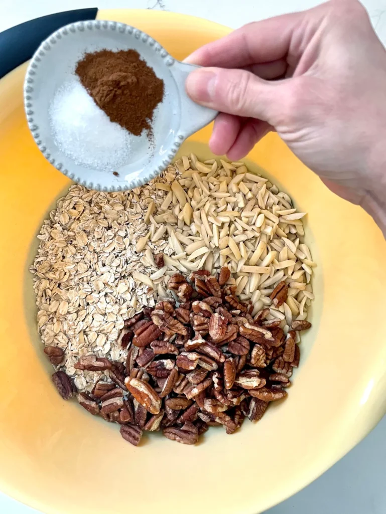 Adding cinnamon and salt to a yellow bowl with oats, pecans, and slivered almonds