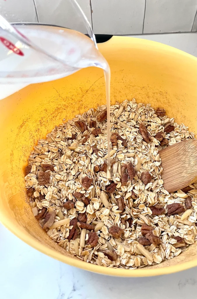 Pouring melted coconut oil into oat and nut mixture in a yellow mixing bowl