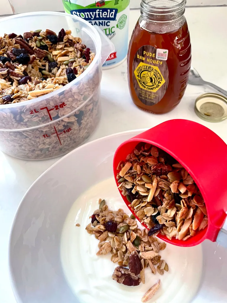 Pouring granola from a red measuring cup over yogurt with container of granola, honey, and yogurt in background