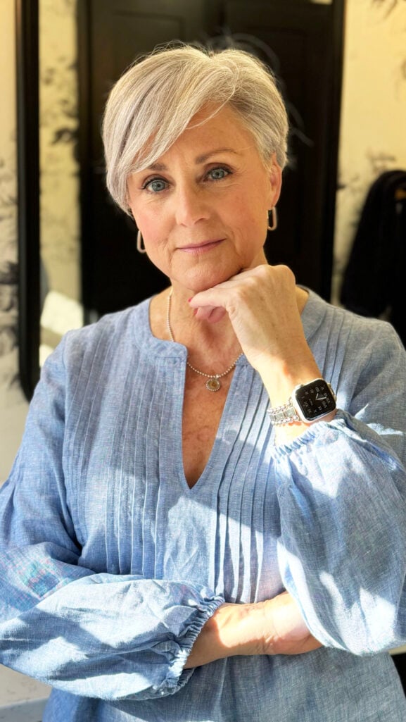 Woman over 50 with short silver hair wearing blue linen tunic and gold jewelry showing healthy glowing skin