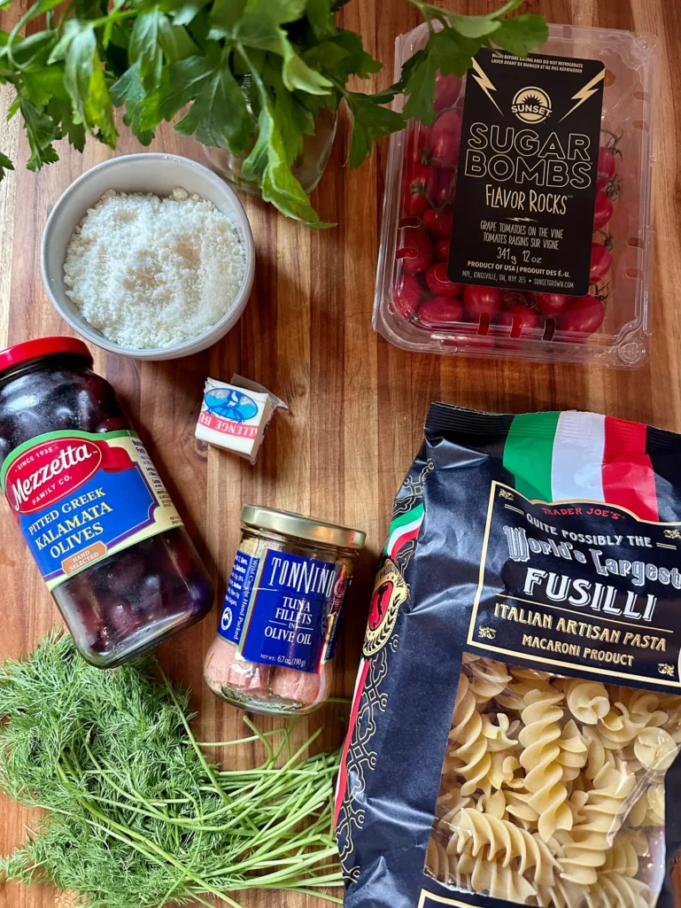 Ingredients for Greek pasta including fusilli, Tonnino tuna, Kalamata olives, grape tomatoes, Parmesan, fresh dill, and parsley on a wooden cutting board