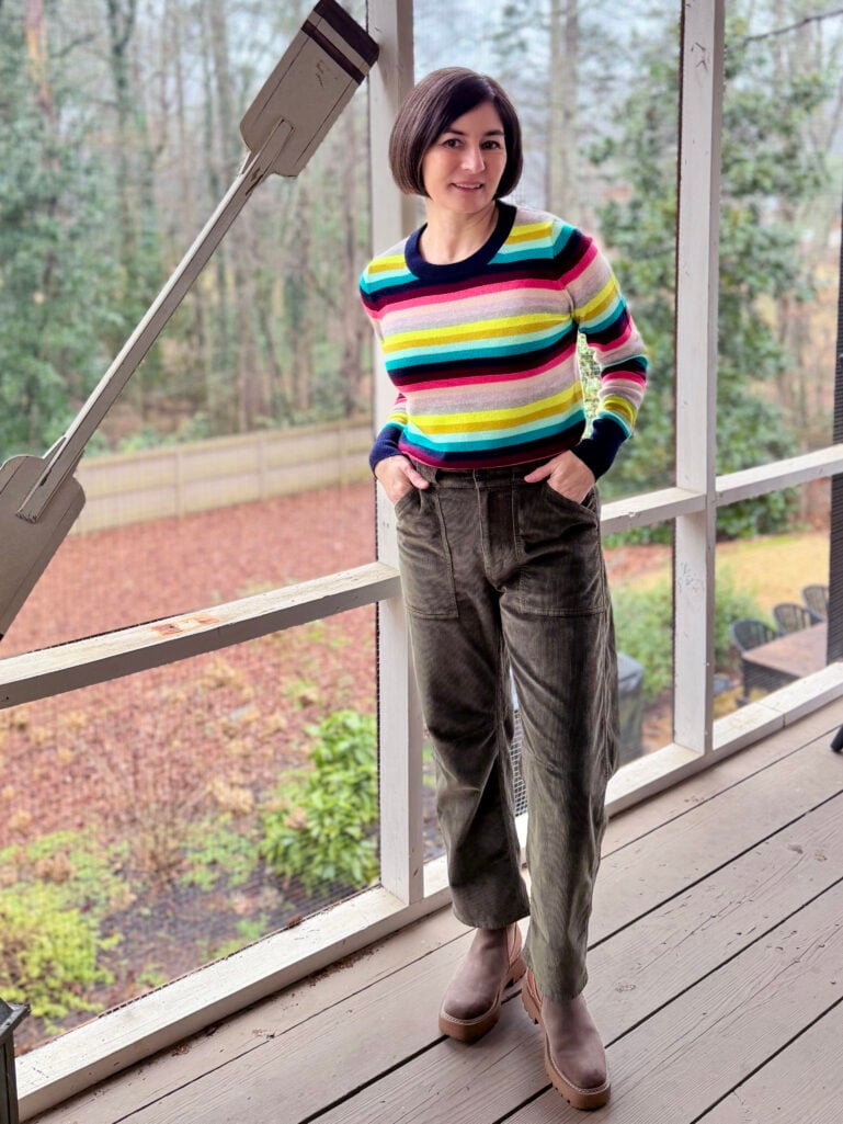 Kelly wearing a colorful rainbow-striped cashmere sweater, olive corduroy barrel-leg pants, and taupe Chelsea boots on the screened porch, hands in pockets