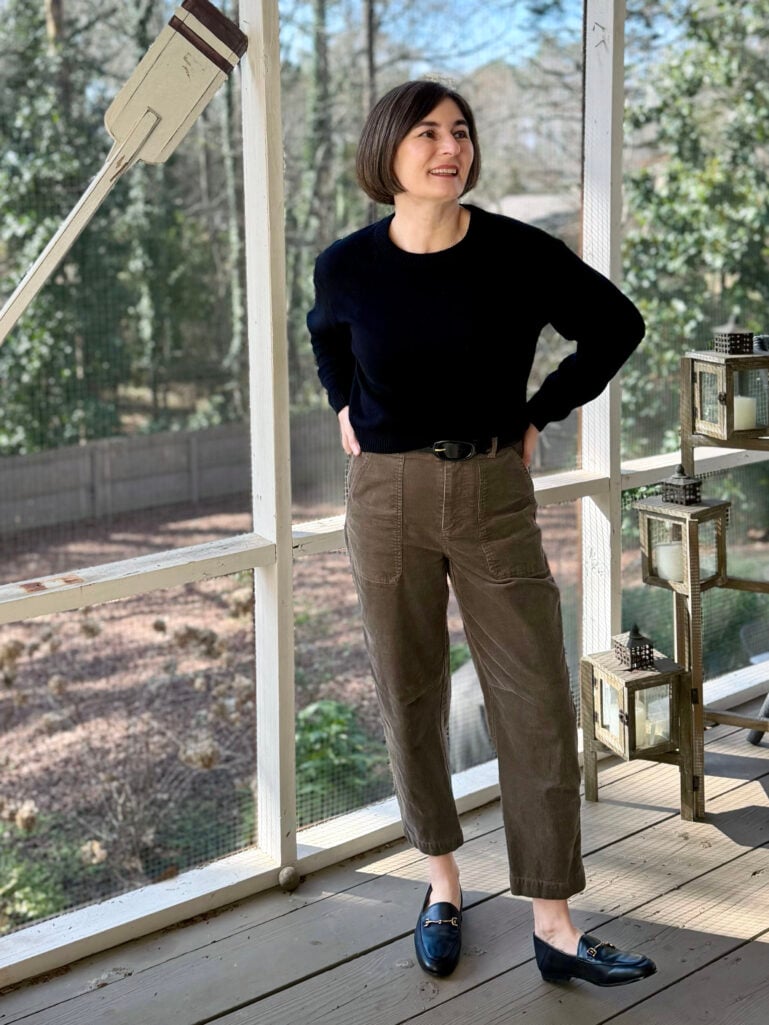 Kelly wearing a black cashmere crewneck sweater, brown barrel leg corduroy pants, black belt, and black Loraine bit loafers on the screened porch