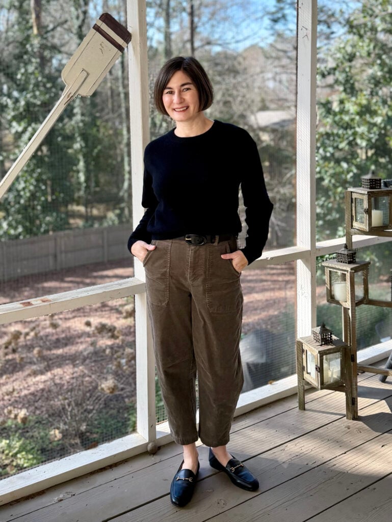 Full-length view of Kelly in her black sweater and brown corduroys, hands in pockets, smiling on the screened porch
