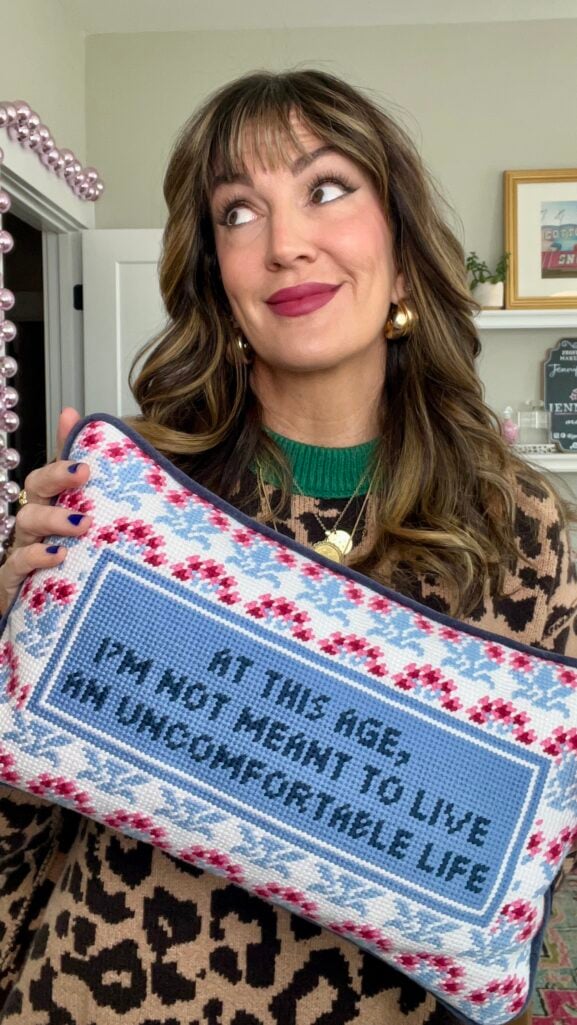 Woman with highlighted brown hair holding Furbish Studio needlepoint pillow reading "At This Age, I'm Not Meant To Live An Uncomfortable Life" in blue with pink floral border, wearing leopard print sweater with green trim, gold hoop earrings, layered gold necklaces, berry lipstick, and royal blue nail polish