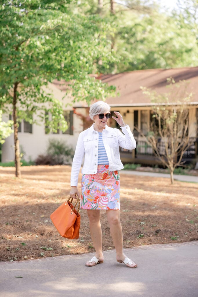 Beth in a blue and white striped tee with a colorful tropical floral skirt, white denim jacket, orange tote bag, and white sandals — mixing stripes and florals for summer