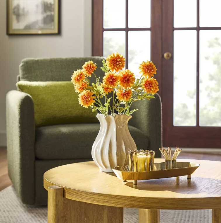 Round wood coffee table styled with a sculptural white ceramic vase filled with orange flowers and a brass tray holding glass votives and matches, set in a cozy living room with a green armchair and French doors in the background.