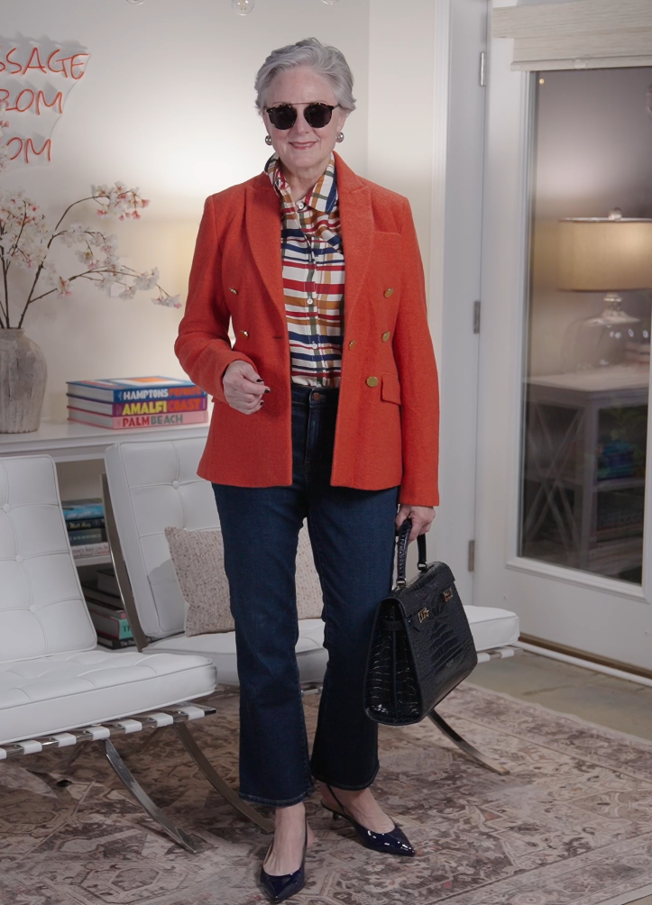 Woman wearing an orange blazer over a multicolor plaid shirt with dark jeans, navy heels, and a structured black handbag.