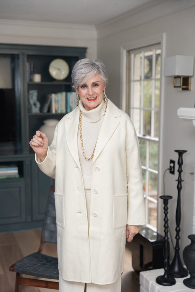 Woman over 50 in cream sleeveless turtleneck sweater with gold chain jewelry styled with cream blazer coat