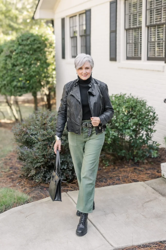 Beth wearing sage green slim wide-leg utility pants with black leather moto jacket, black ribbed turtleneck, studded belt, black combat boots, and tooled leather shoulder bag