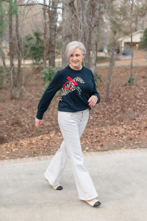 Woman with short gray hair wearing a navy sweater with zebra motif and red bow detail, paired with white straight-leg pants and black cap-toe ballet flats, walking along a wooded path.