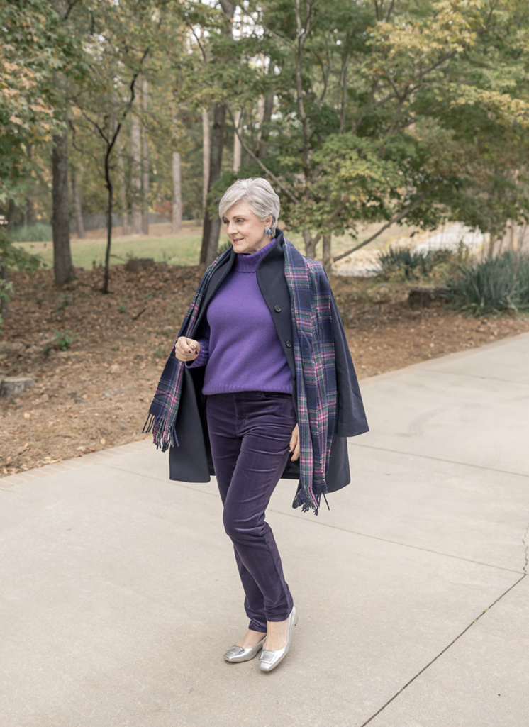 Woman wearing a purple turtleneck sweater with plum velvet pants, layered under a navy coat and plaid scarf, walking outdoors on a paved path with autumn trees in the background.